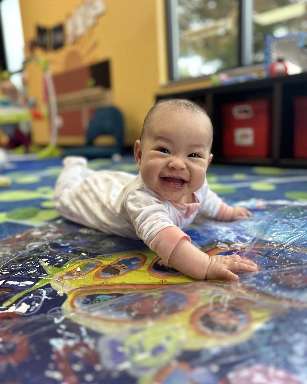 A smiling infant crawling across a colorful activity mat in the infant classroom