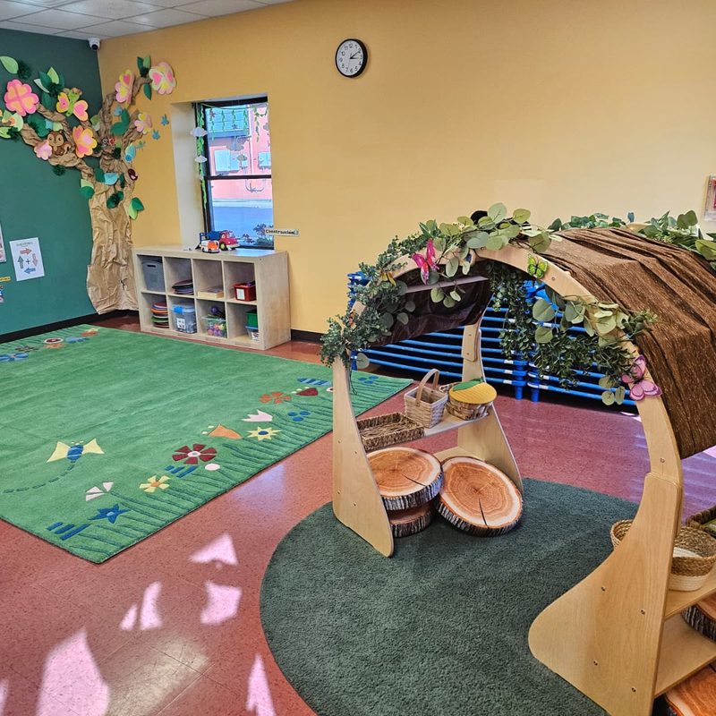 A preschool classroom with a paper-flower tree mural, a green rug and a leafy reading nook