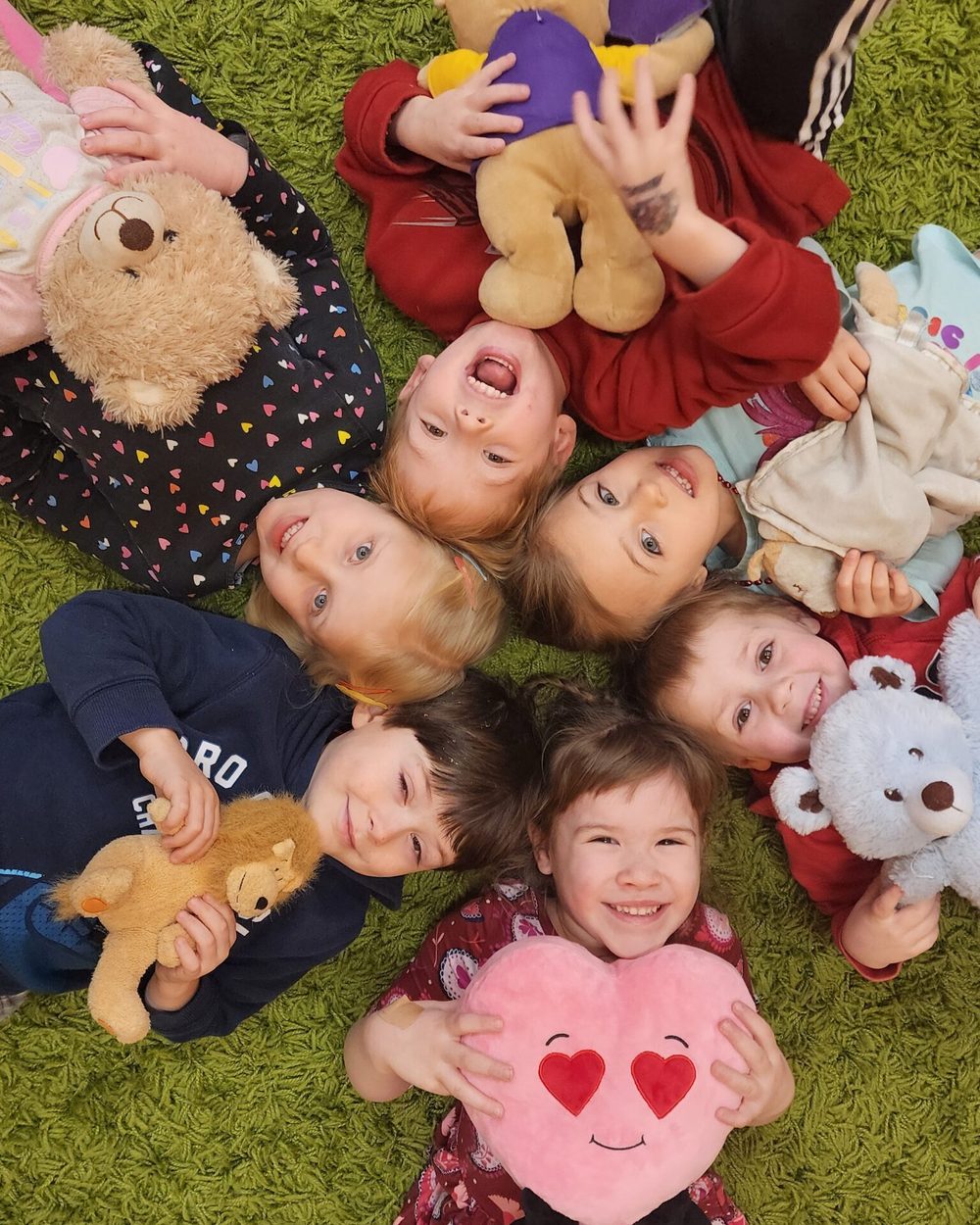 Preschoolers lying in a circle on a green rug, heads together, each hugging a stuffed animal