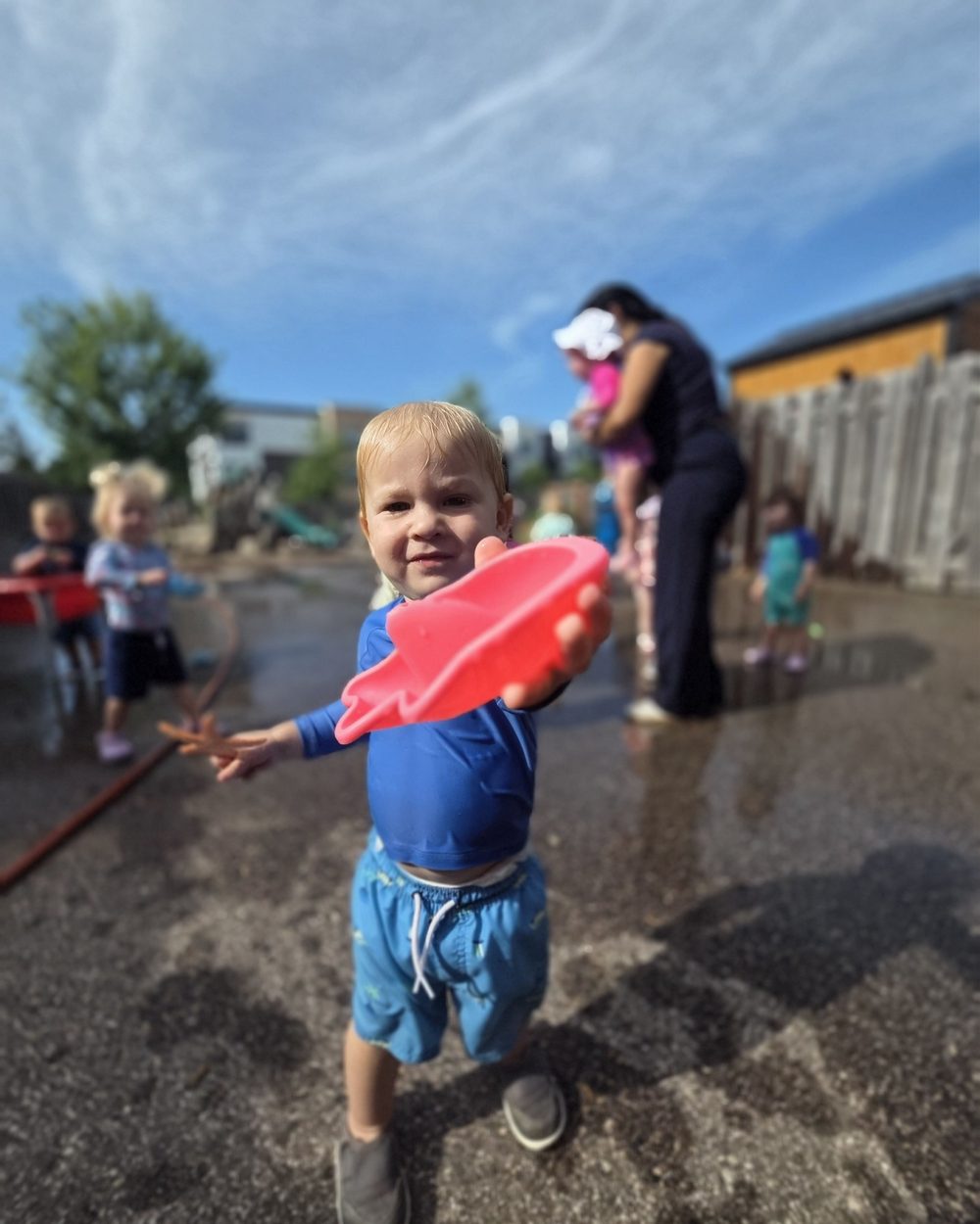 A toddler holding up a red water toy in the splash yard on a summer day