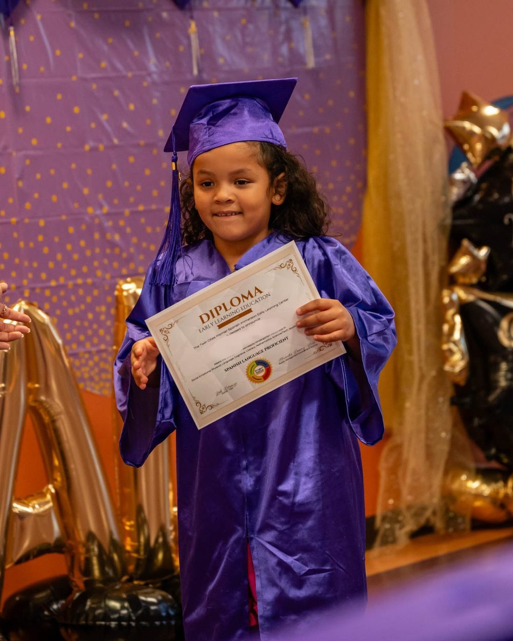 A Pre-K graduate in a purple cap and gown holding a diploma that reads Spanish Language Proficient