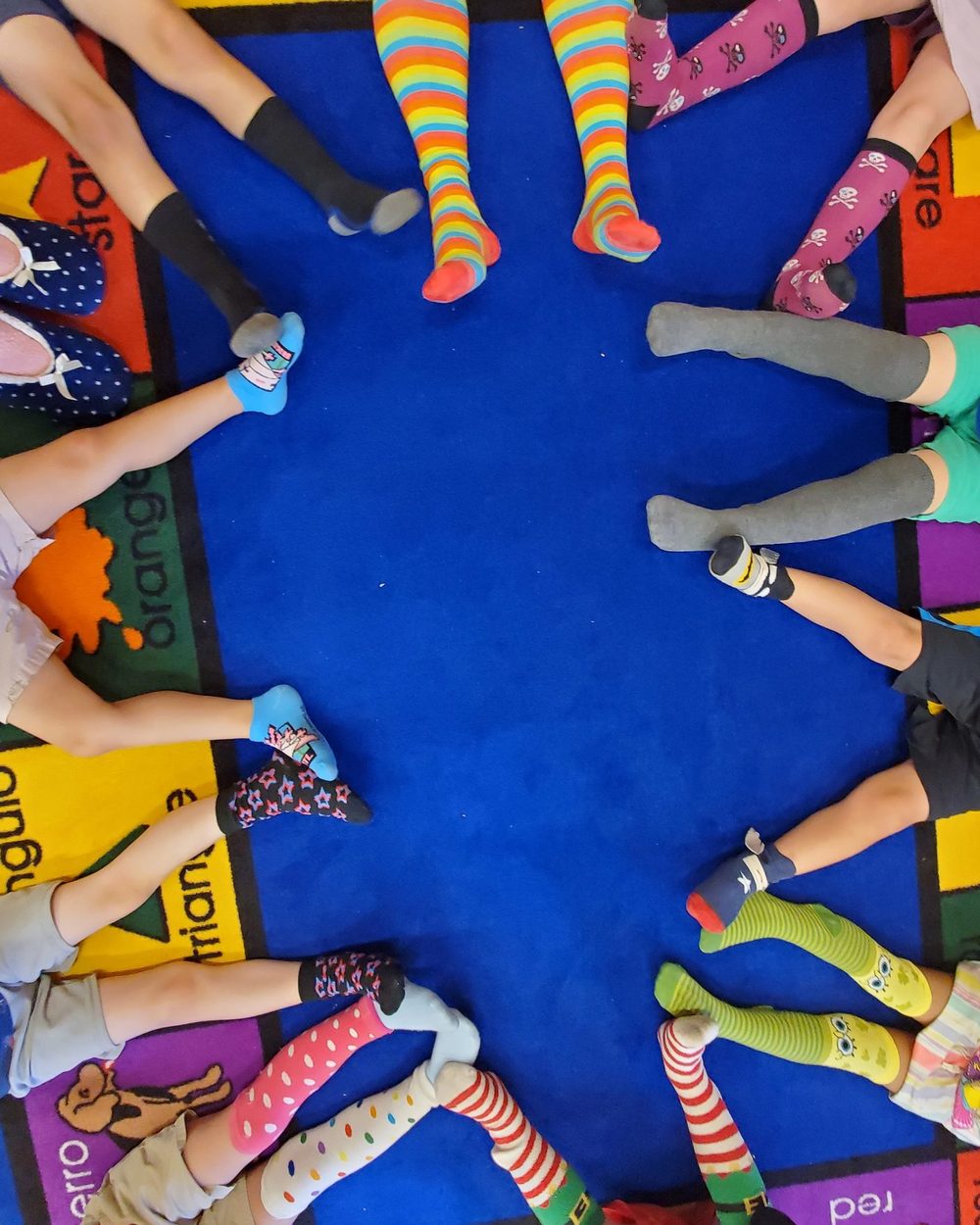 Children's stocking feet in a circle on a classroom rug printed with Spanish and English color words