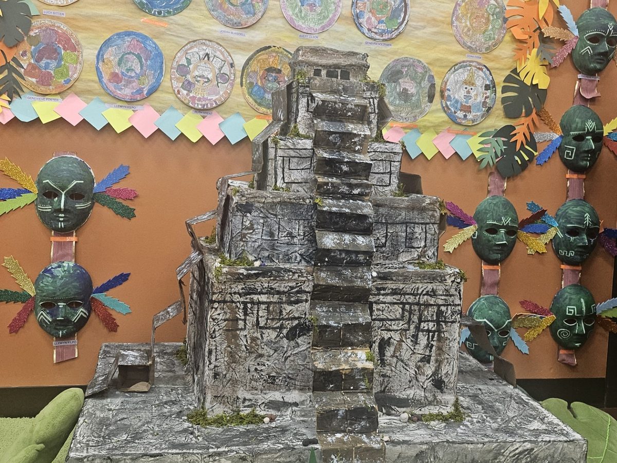 A student-built model of the Chichén Itzá pyramid in front of a wall of Aztec calendar art