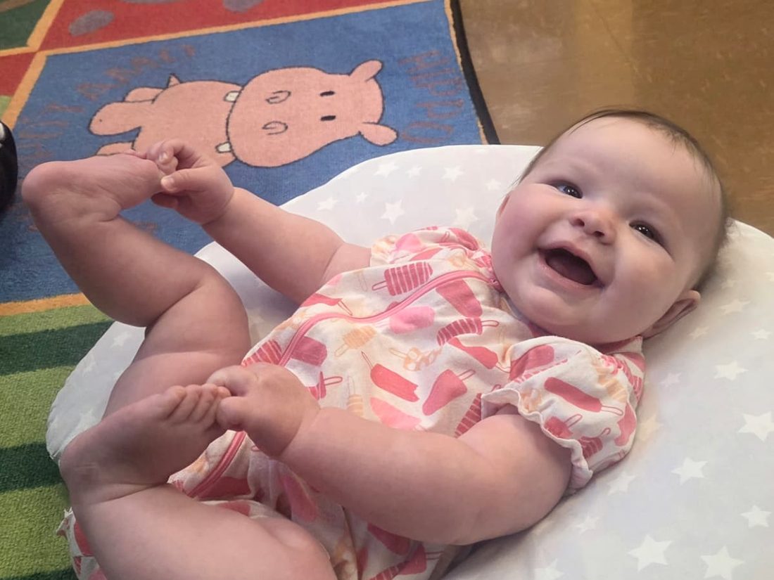 A smiling infant lying on a pillow on the classroom rug, feet in the air
