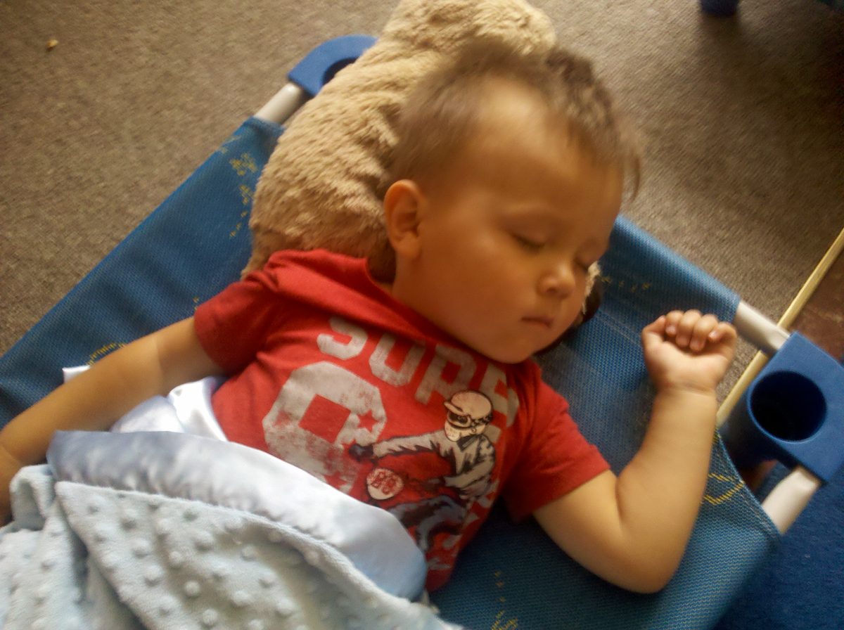 A sleeping toddler lying on a blue cot in a classroom with a teddy bear and a soft blanket