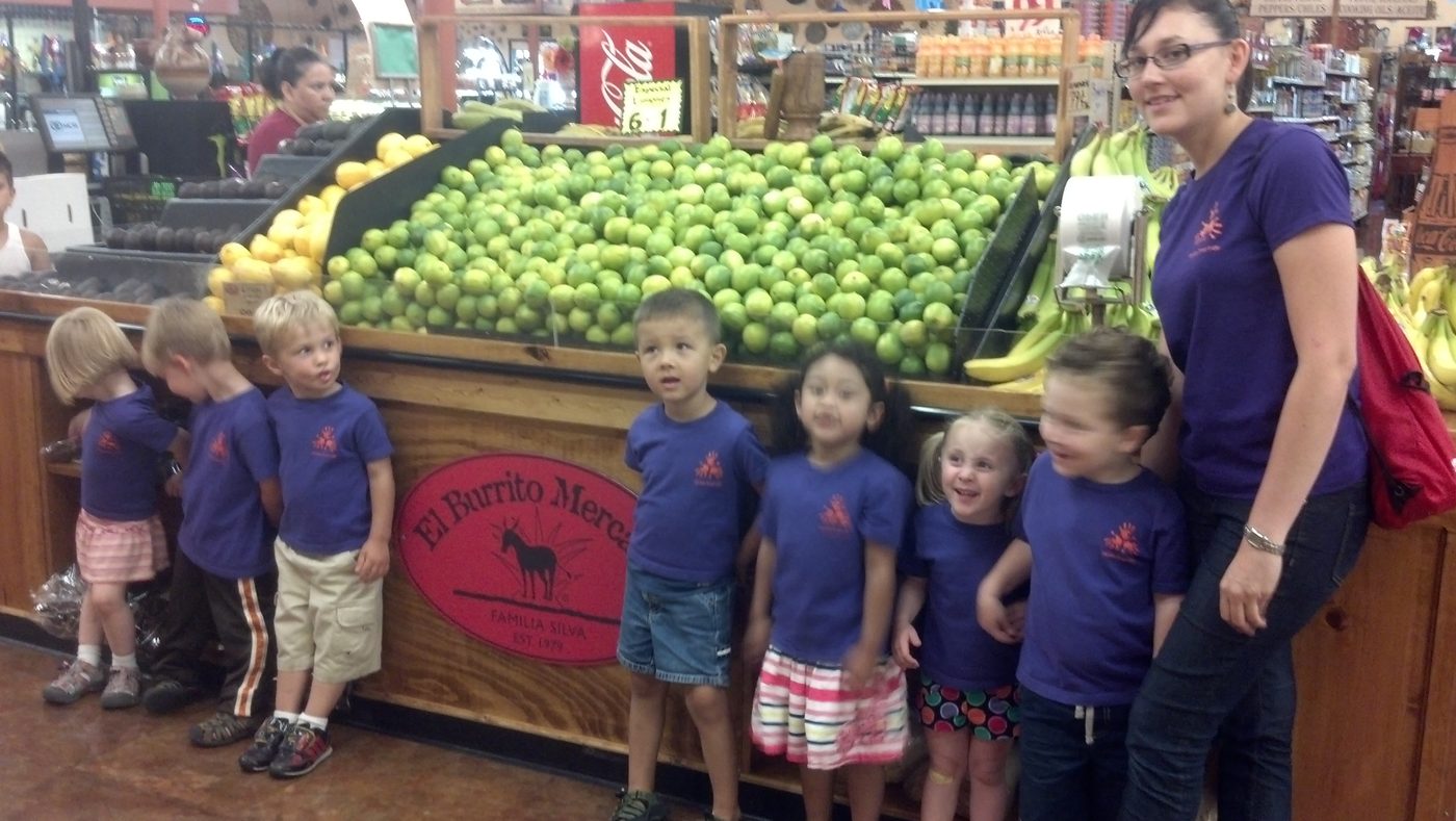 A line of young children in matching purple Center t-shirts standing with a teacher in front of a large display of limes and lemons at a grocery market