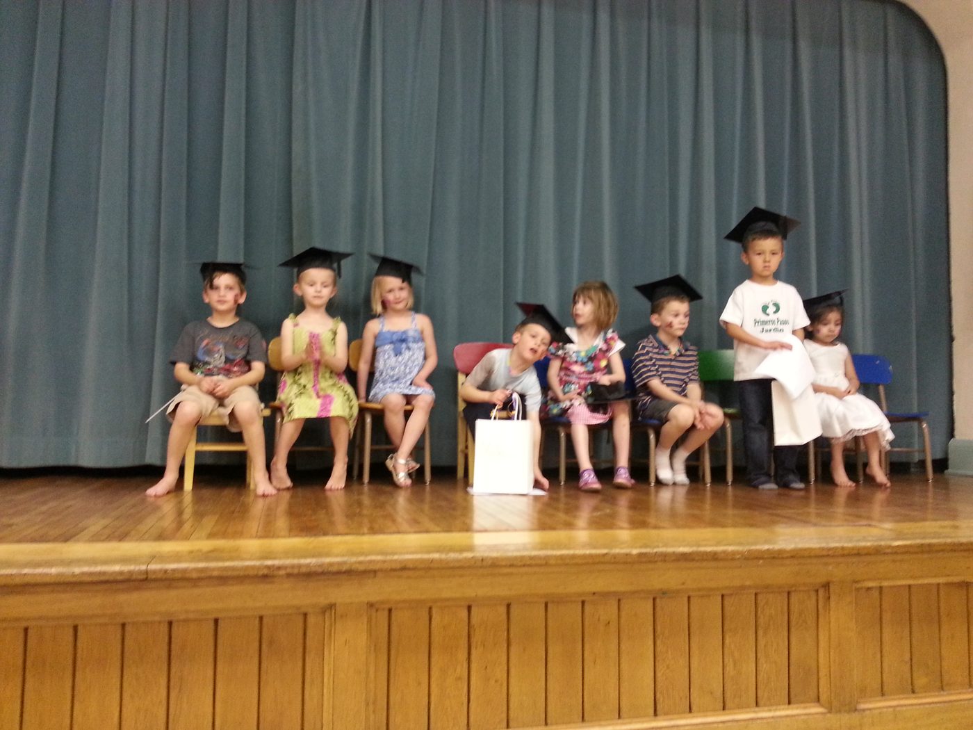 Pre-K graduates in black mortarboard caps seated on small chairs on a wooden stage in front of a blue-grey curtain, one standing to read aloud