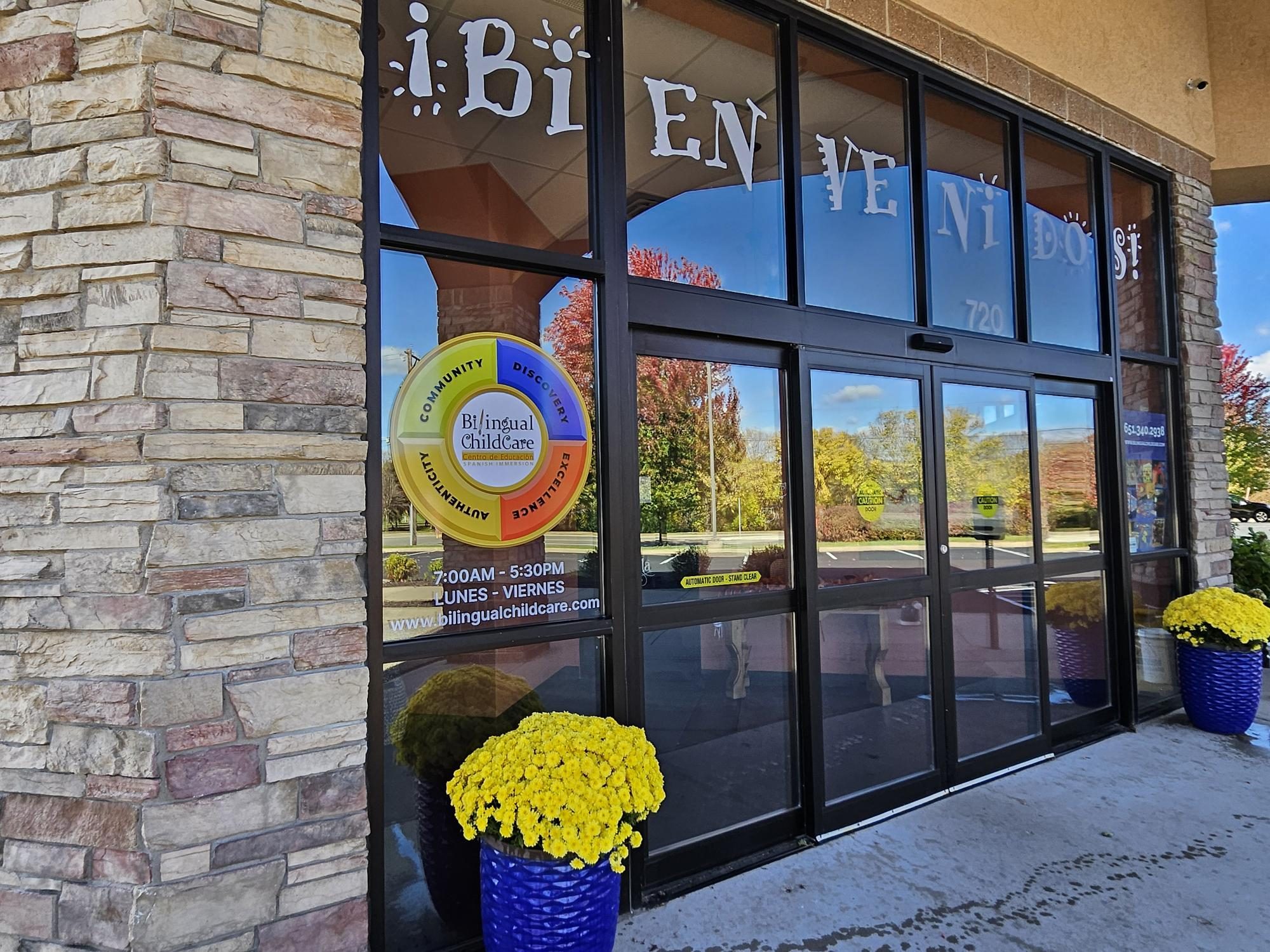 The Mahtomedi center entrance at 720 Wildwood Road, with a ¡Bienvenidos! decal across the windows and planters of yellow mums