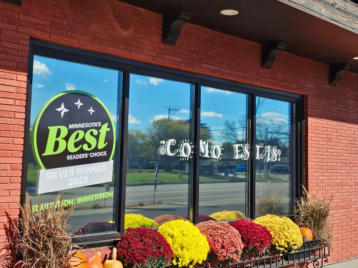 The Roseville storefront on County Road B, with a ¿Cómo estás? greeting across the window and fall mums along the sill